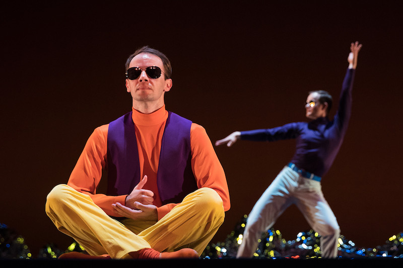 A man in sunglasses sits cross-legged in the foreground. A dancer in a purple turtleneck poses in the background.